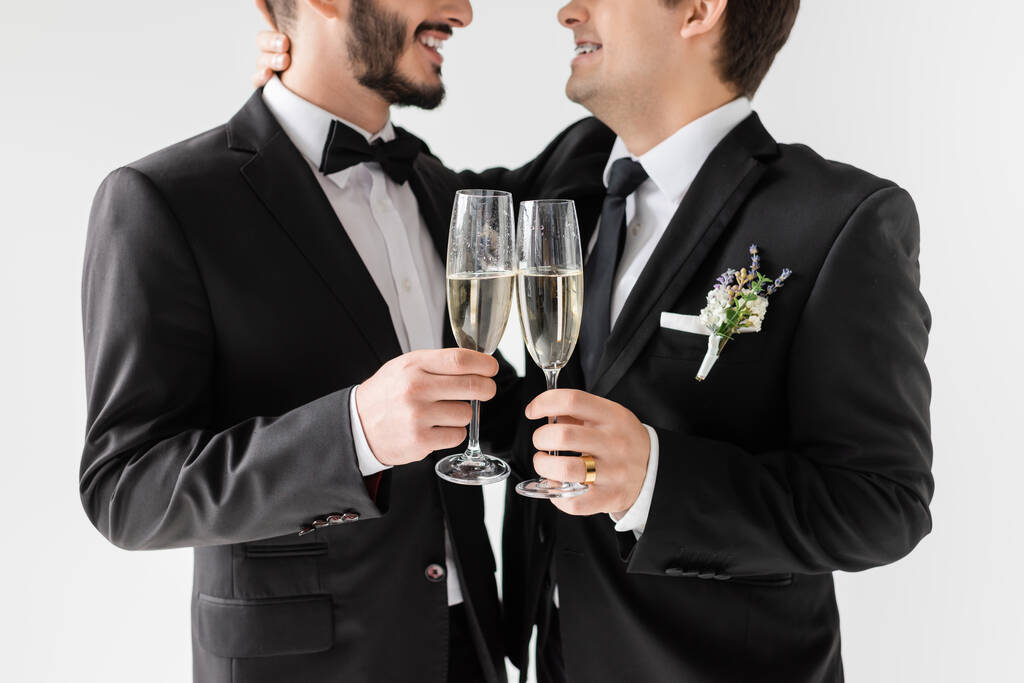 Oříznutý pohled na usmívajícího se homosexuálního ženicha v rovnátkách a obleku s boutonniere objímání vousatý přítel a opékání se šampaňským během svatebního obřadu izolované na šedé  - Fotografie, Obrázek