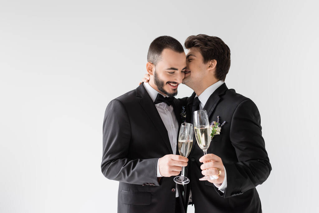Young homosexual man in suit with boutonniere whispering in ear of smiling boyfriend with glass of champagne during wedding ceremony isolated on grey  - Photo, Image