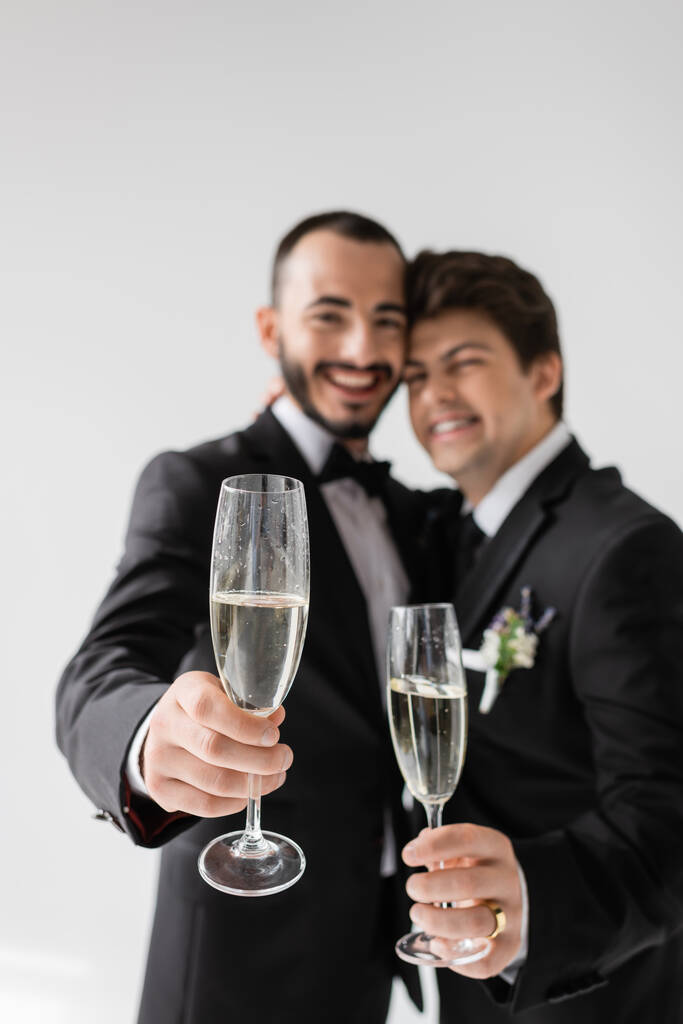 Blurred lgbt couple in elegant formal wear smiling and showing champagne glasses at camera during wedding celebration isolated on grey  - Photo, Image