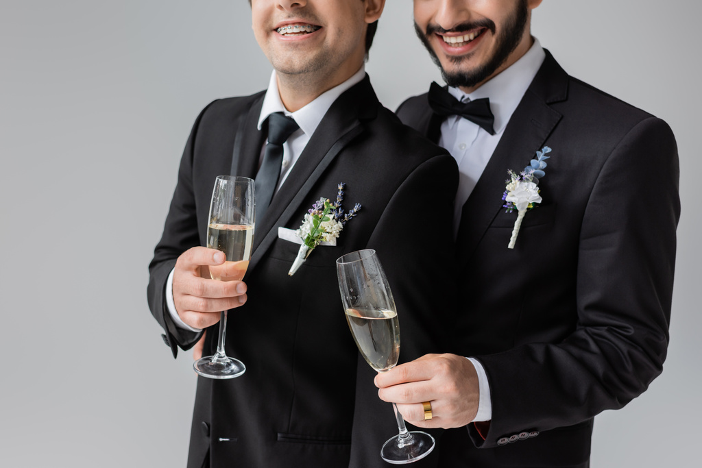 Vista cortada de noivos homossexuais sorridentes em traje clássico com boutonnieres florais segurando copos de champanhe enquanto celebra casamento isolado em cinza  - Foto, Imagem