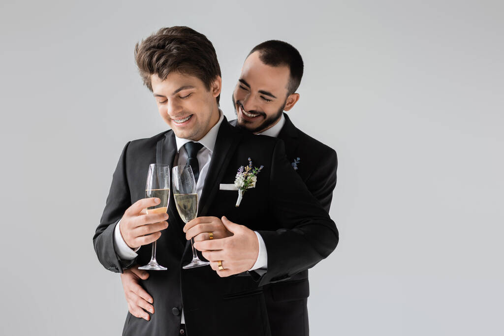 Χαμογελαστός και γενειοφόρος ομοφυλόφιλος γαμπρός αγγίζοντας το χέρι του κομψού φίλου στο κοστούμι κρατώντας ποτήρια σαμπάνιας κατά τη διάρκεια της γαμήλιας γιορτής απομονώνονται σε γκρι  - Φωτογραφία, εικόνα