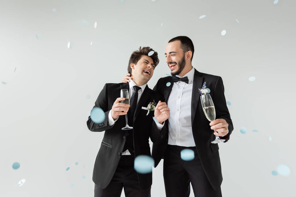 Fröhlicher homosexueller Bräutigam im eleganten Anzug umarmt jungen Freund mit einem Glas Champagner, während er unter fallendem Konfetti auf grauem Hintergrund die Ehe feiert - Foto, Bild