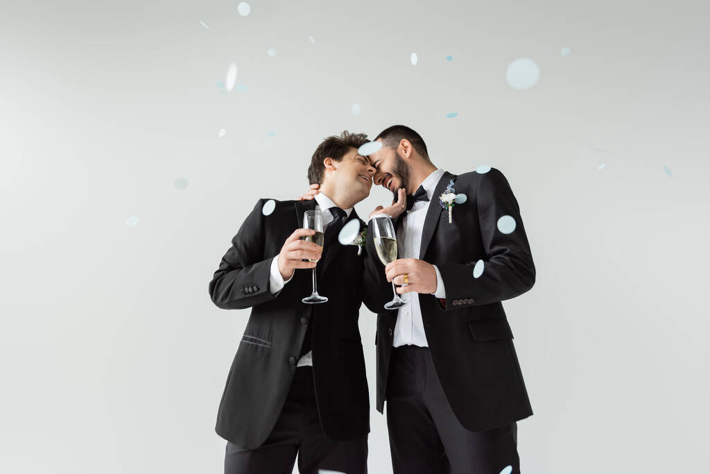 Alegres novios homosexuales en trajes que se tocan mientras sostienen copas de champán y celebran el matrimonio bajo la caída de confeti sobre fondo gris - Foto, Imagen
