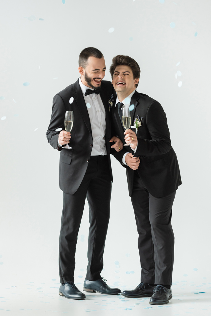 Longitud completa de novios del mismo sexo muy alegres en trajes que sostienen champán y se divierten bajo la caída de confeti festivo durante el día de su boda sobre fondo gris - Foto, Imagen