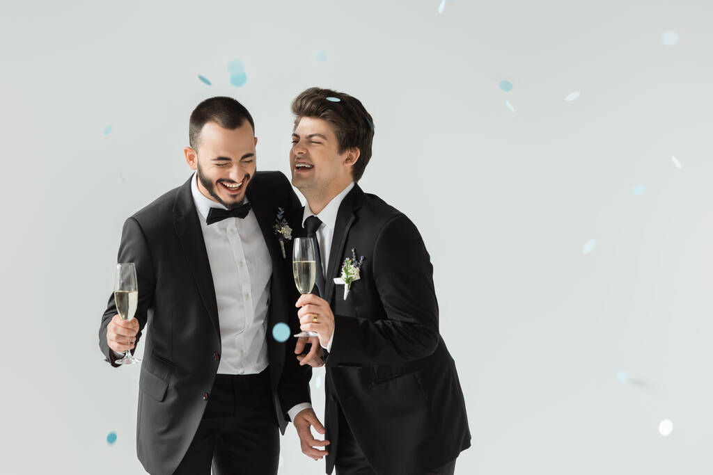 Vrolijke gay bruidegom holding champagne in de buurt elegant vriendje in klassiek pak terwijl staan onder vallende confetti tijdens de bruiloft op grijze achtergrond - Foto, afbeelding