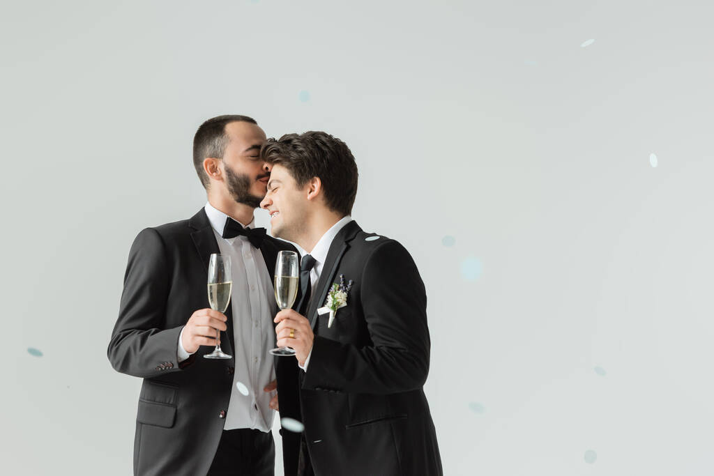 Der bärtige homosexuelle Bräutigam in offizieller Kleidung hält ein Glas Champagner in der Hand und küsst seinen lächelnden jungen Freund unter Konfetti während der Hochzeitszeremonie auf grauem Hintergrund - Foto, Bild