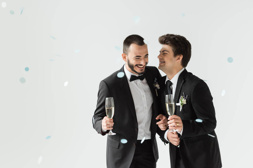 Positive same sex grooms in classic attire holding champagne while standing under falling confetti during wedding ceremony on grey background - Photo, Image