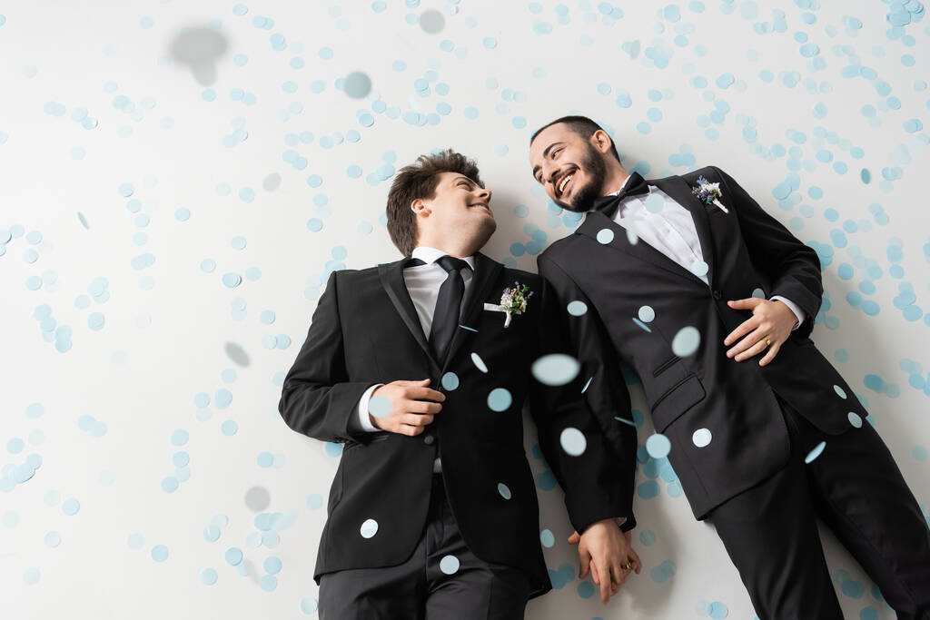 Bovenaanzicht van positieve bruidegoms van hetzelfde geslacht in formele slijtage die elkaar aankijken en elkaars hand vasthouden terwijl ze onder vallende confetti op grijze achtergrond liggen  - Foto, afbeelding