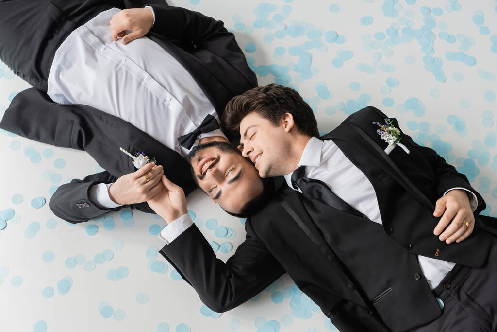 Top view of smiling gay groom touching hand of bearded boyfriend in classic suit lying together on festive confetti during wedding celebration on grey background  - Photo, Image