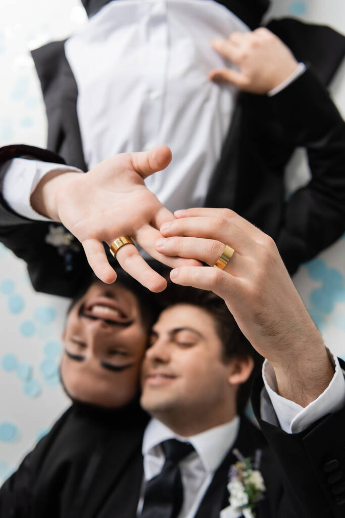 Vista dall'alto di sposi sfocati dello stesso sesso in abiti che toccano le dita l'uno dell'altro in fedi nuziali mentre giacciono su coriandoli festivi su sfondo grigio  - Foto, immagini
