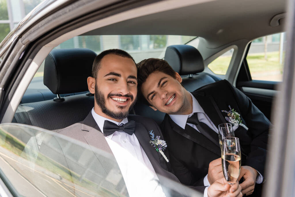 Young and positive newlyweds in formal wear with floral boutonnieres holding glasses of champagne and looking at camera from car window while sitting on backseat  - Photo, Image
