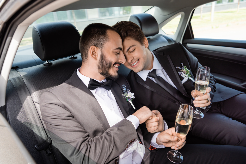 Sorrindo jovens noivos gays em ternos elegantes fechando os olhos enquanto segurando as mãos e copos de champanhe enquanto sentado no banco de trás durante a viagem de lua de mel no carro  - Foto, Imagem
