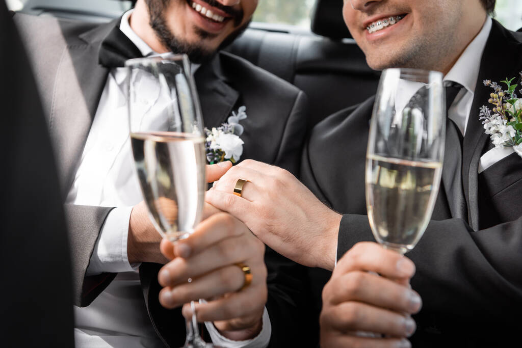 Cropped view of positive same sex newlyweds in classic suits with boutonnieres holding hands and blurred glasses of champagne during road trip in car  - Photo, Image