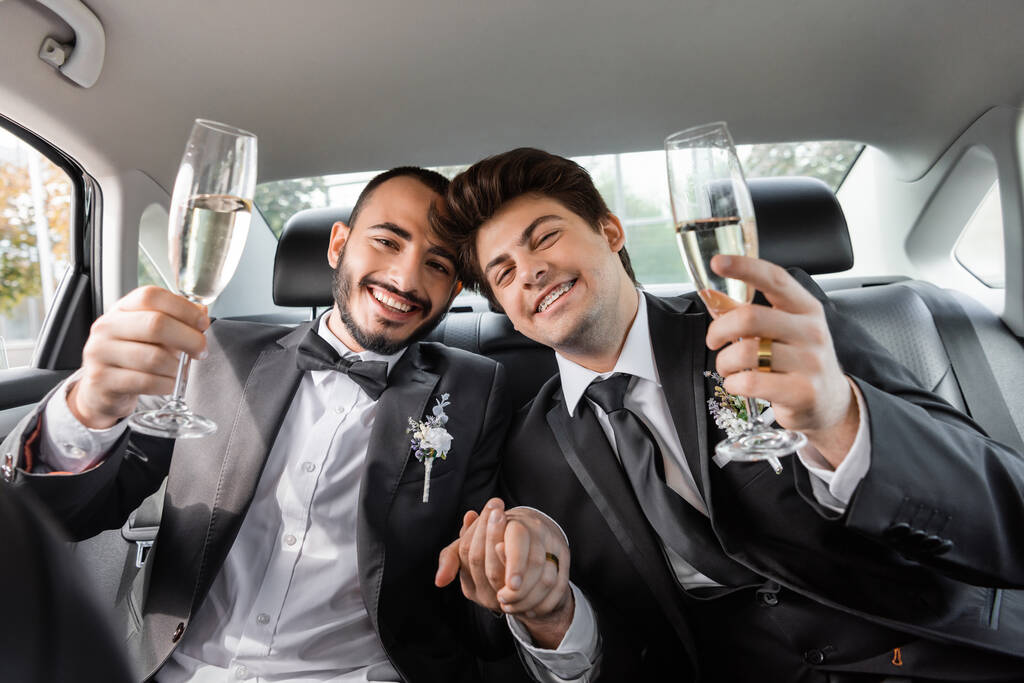 Lächelnde junge schwule Frischvermählte in formeller Kleidung, die Hände und Champagner halten, während sie auf dem Rücksitz des Autos in die Kamera schauen  - Foto, Bild
