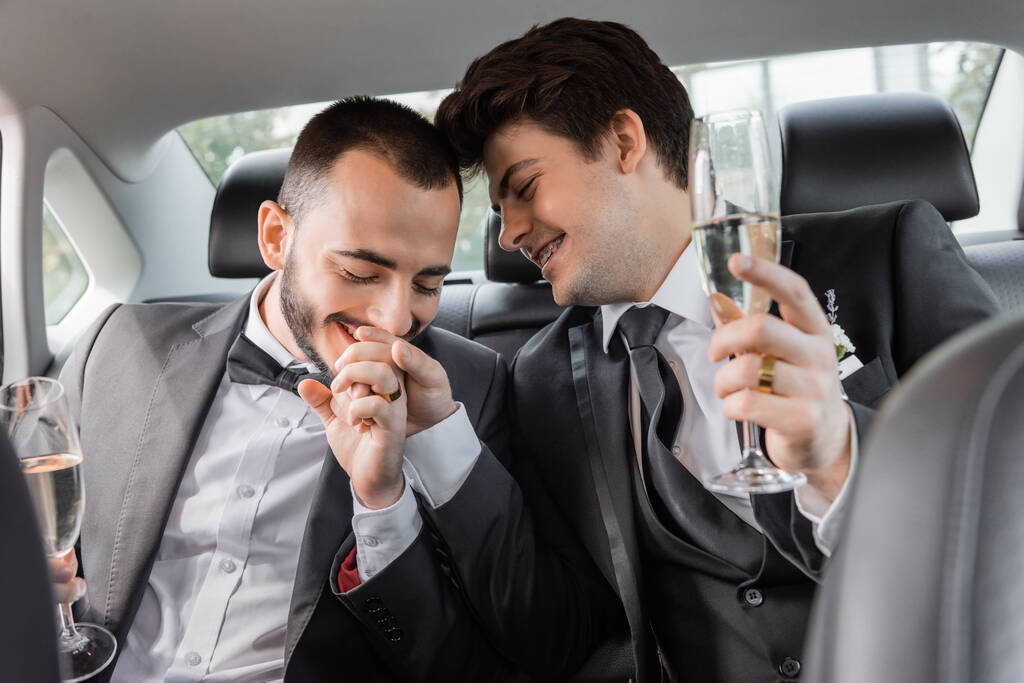 Positive gay groom in elegant formal wear kissing hand of young partner in braces and holding champagne glass during honeymoon road trip and sitting sitting on backseat of car  - Photo, Image