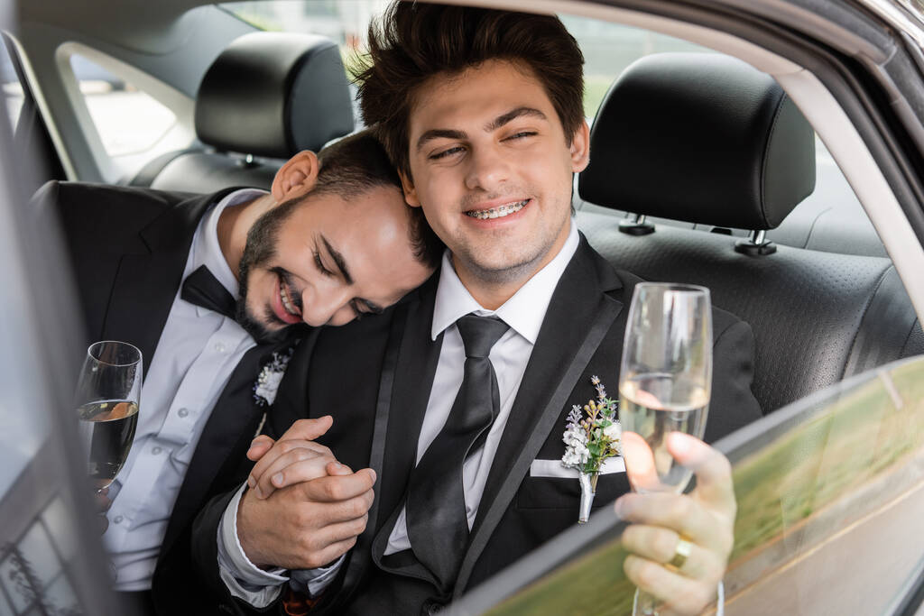 Χαμογελαστός και νεαρός gay γαμπρός με τιράντες σε κομψό κοστούμι με μπουτονιέρα κρατώντας ένα ποτήρι σαμπάνια και το χέρι του φίλου, ενώ κάθεται στο πίσω κάθισμα του αυτοκινήτου κατά τη διάρκεια του μέλιτος  - Φωτογραφία, εικόνα