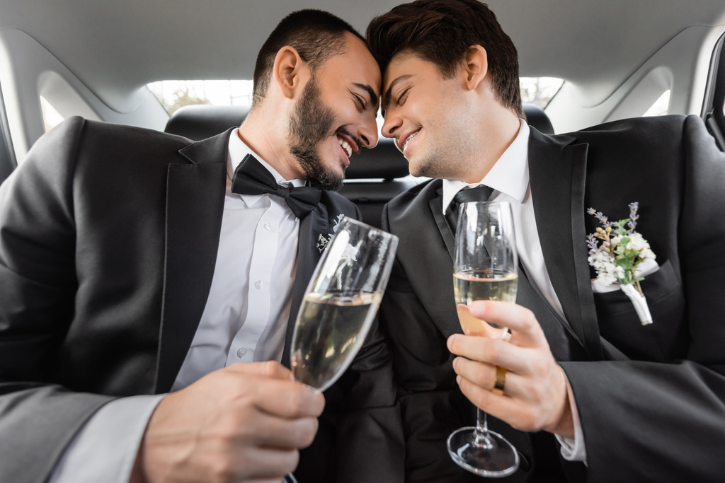 uśmiechnięty i młody gej panny młode w formalny odzież z butonierki siedzi nos w nos i trzymając szampan na tylnym siedzeniu samochód po wesele uroczystości - Zdjęcie, obraz