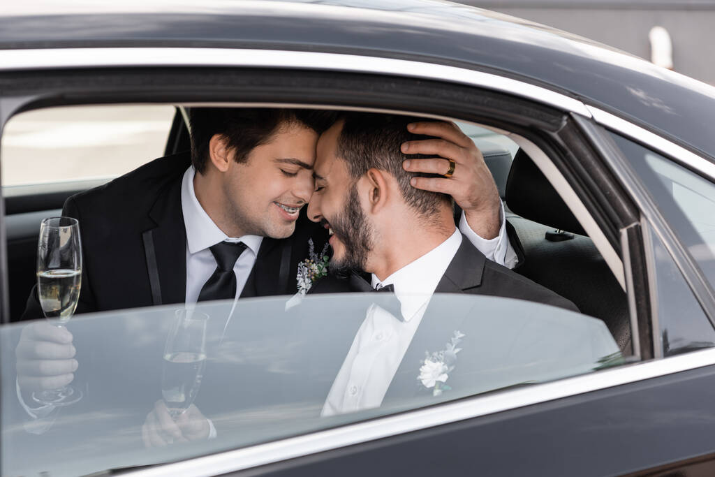 Young homosexual groom in braces and formal wear holding champagne and hugging cheerful bearded boyfriend while while sitting on backseat of car before going to honeymoon  - Photo, Image