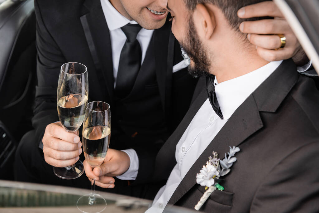 Mosolygó homoszexuális vőlegény formális viselet kezében pezsgős üveg és ölelkező szakállas barátja, miközben ünnepli esküvő autóban nászúton  - Fotó, kép