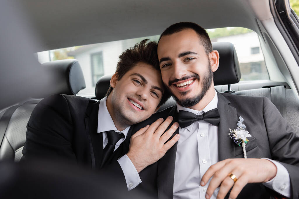 Cheerful homosexual groom with braces in suit hugging bearded boyfriend and looking at camera after wedding celebration while going on honeymoon  - 写真・画像