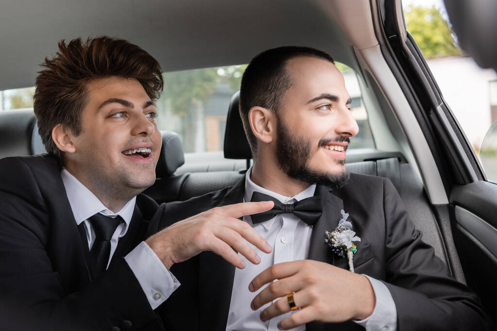Joyeux marié gay dans des bretelles et costume élégant pointant du doigt près de copain barbu et regardant ensemble par la fenêtre de la voiture après la célébration du mariage tout en allant en lune de miel  - Photo, image