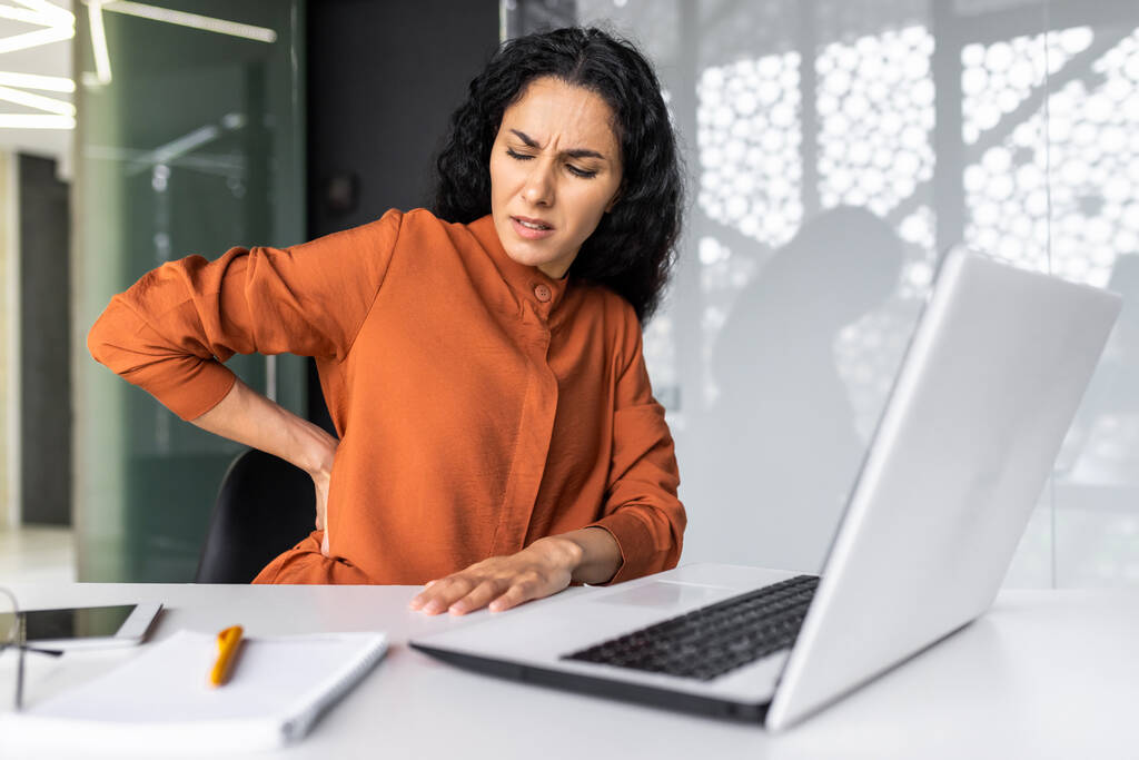 Υπερκόπωση ισπανόφωνη γυναίκα που εργάζεται μέχρι αργά, επιχειρηματίας που έχει σοβαρό πόνο στην πλάτη, γυναίκα εργαζόμενη μέσα στο γραφείο κάθεται στο τραπέζι χρησιμοποιώντας φορητό υπολογιστή στη δουλειά - Φωτογραφία, εικόνα