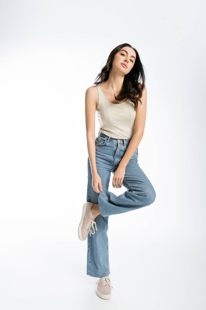 full length of alluring woman with shiny brunette hair, natural makeup and perfect skin posing in tank top and touching denim jeans on white background - Photo, Image