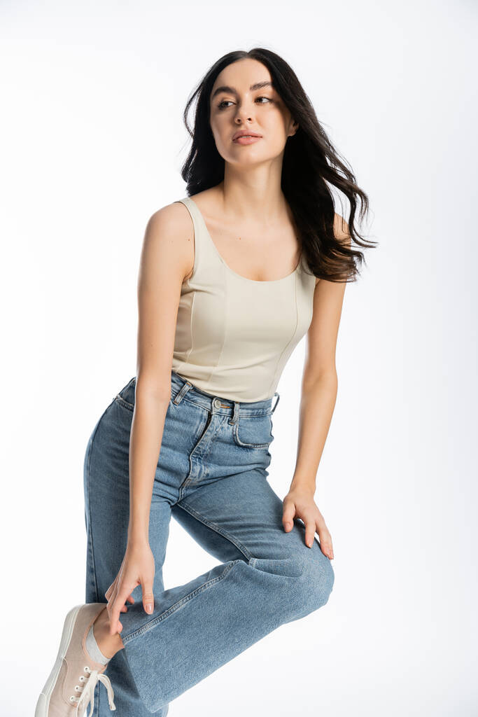 gorgeous and young woman with shiny brunette hair, natural makeup and perfect skin posing in tank top and touching denim jeans while looking away on white background - Photo, Image