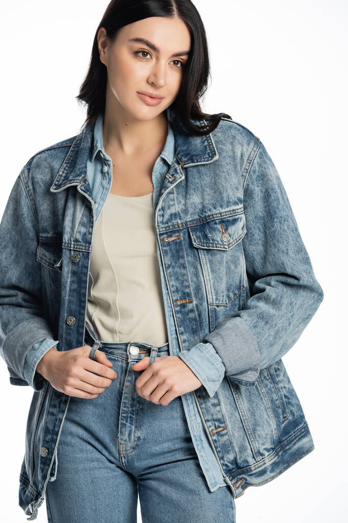 young and fashionable model with brunette hair and natural makeup posing in stylish denim outfit while standing and looking away on white background - Photo, Image