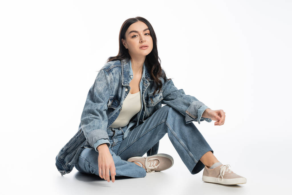 full length of chic young model with brunette hair and natural makeup posing in trendy denim outfit while sitting and looking at camera on white background - Photo, Image