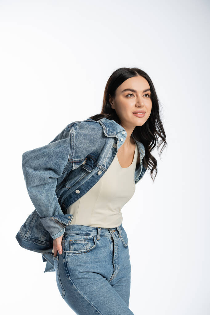 dreamy young woman with gorgeous brunette hair posing with hand in pocket of blue jeans while standing in denim jacket and looking away on white background - Photo, Image