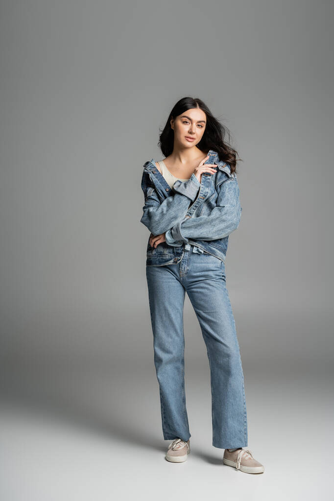 full length of charming woman with brunette hair posing while standing in stylish blue jeans and trendy denim jacket and looking at camera on grey background - Photo, Image