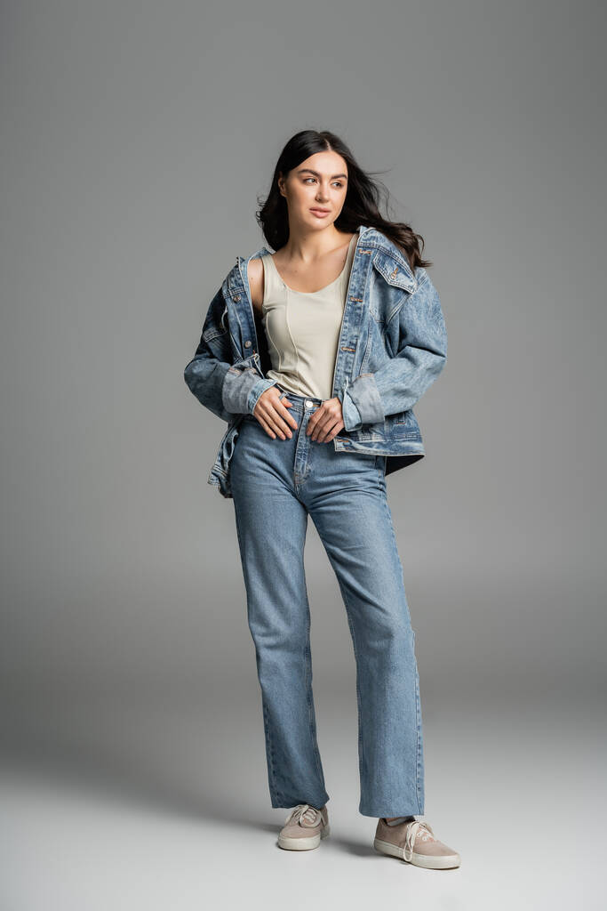 повна довжина чарівної молодої моделі з брюнеткою волосся позує, стоячи в стильних блакитних джинсах і джинсовій куртці і дивлячись на сірий фон
  - Фото, зображення
