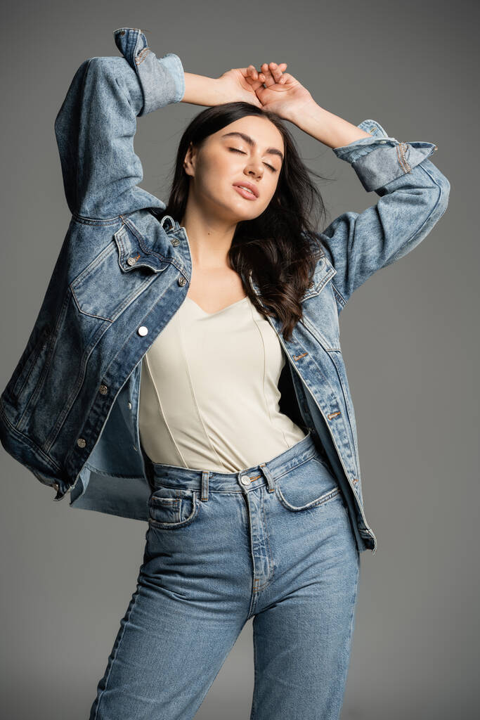 chic young woman with gorgeous hair posing with hands above head and closed eyes while standing in stylish blue jeans and denim jacket on grey background - Photo, Image
