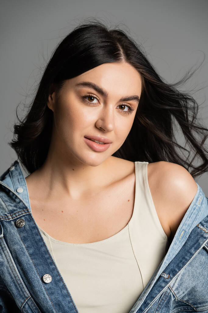 portrait of charming young woman with brunette hair posing in fashionable and blue denim jacket and looking at camera isolated on grey background - Photo, Image