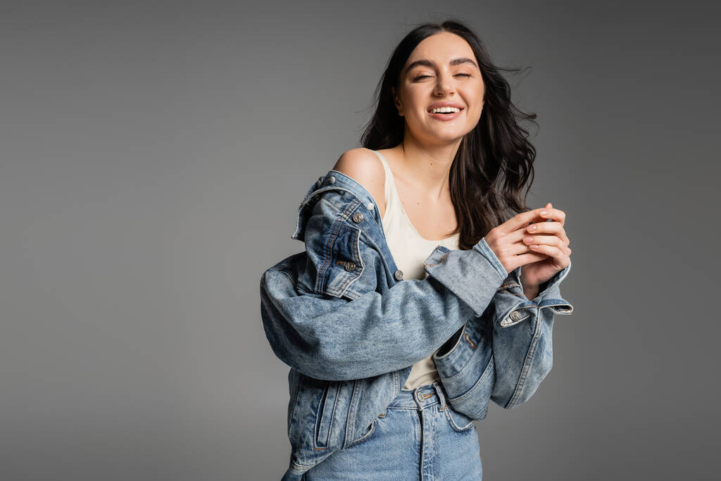 giovane donna positiva con capelli castani in piedi in jeans blu e giacca di jeans alla moda mentre sorride e posa isolata su sfondo grigio - Foto, immagini