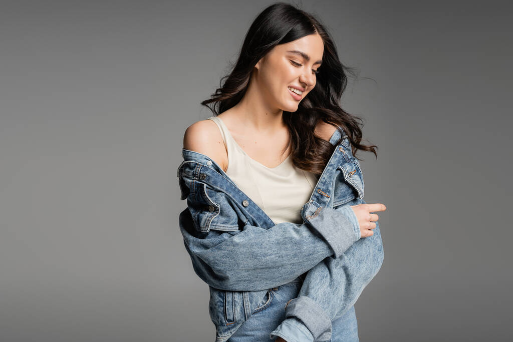 alluring young woman with brunette hair and natural makeup standing in blue jeans and fashionable denim jacket while smiling and posing isolated on grey background - Photo, Image