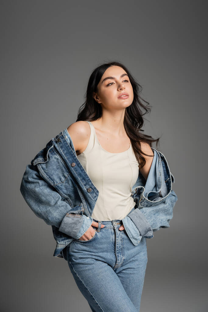 brunette young woman with shiny hair standing with hands in pockets of blue jeans and posing in stylish denim jacket while looking away isolated on grey background - Photo, Image