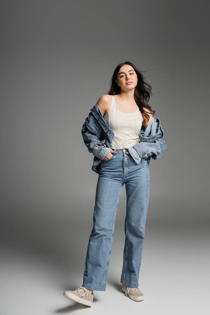full length of young appealing woman with long brunette hair posing in stylish blue jeans and denim jacket while standing with hands in pockets on grey background - Photo, Image