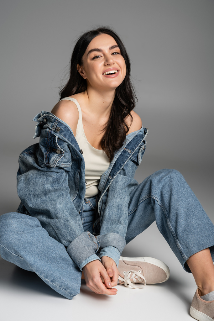 full length of happy woman with long brunette hair and flawless natural makeup posing in stylish blue jeans and denim jacket while sitting on grey background - Photo, Image