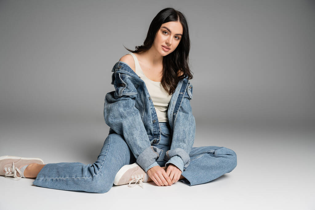 full length of young alluring woman with long brunette hair and flawless natural makeup posing in stylish blue jeans and denim jacket while sitting on grey background - Photo, Image