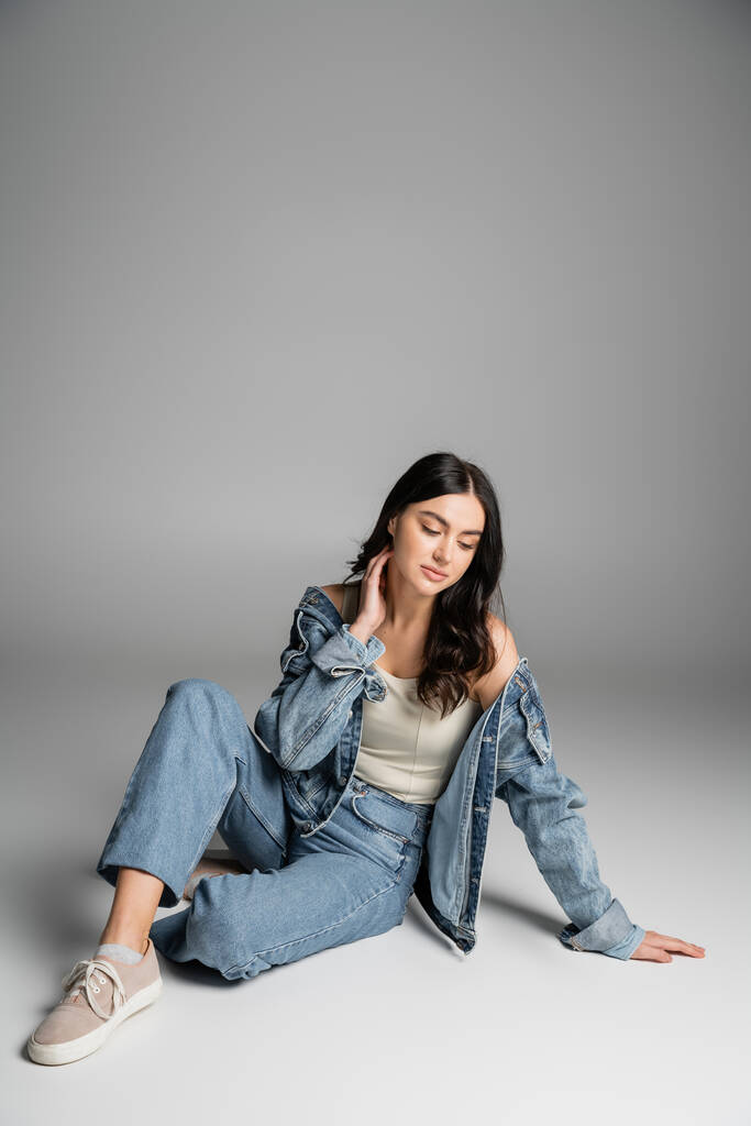 full length of dreamy young woman with long brunette hair and flawless natural makeup posing in stylish blue jeans and denim jacket while sitting on grey background - Photo, Image