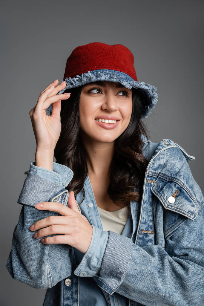 portret van lachende jonge vrouw met vlekkeloze natuurlijke make-up poseren in panama hoed en spijkerjasje terwijl weg te kijken op grijze achtergrond - Foto, afbeelding