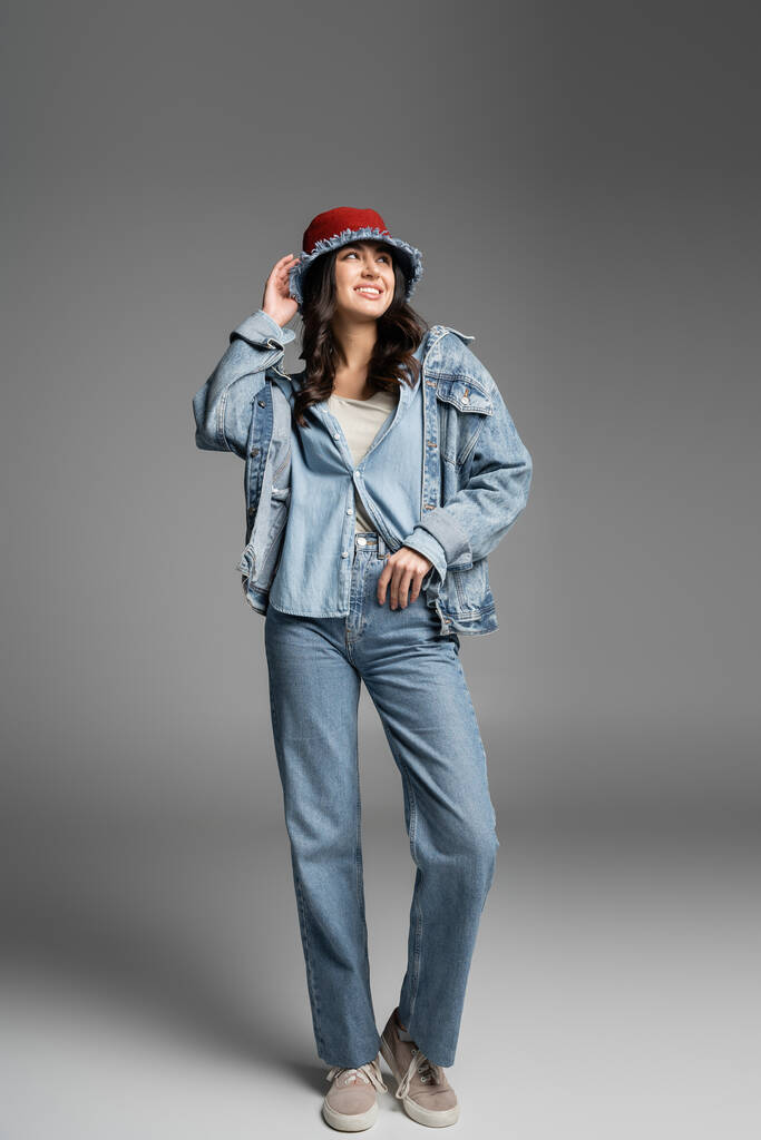 full length of young happy woman with flawless natural makeup adjusting denim panama hat and posing in blue jeans and jacket while standing and looking away on grey background - Photo, Image