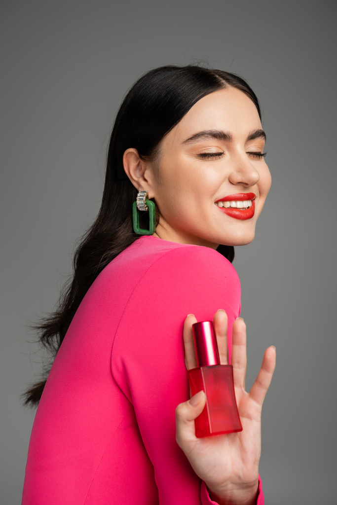 tevreden vrouw met brunette haar, trendy oorbellen, rode lippen en stijlvolle magenta jurk holding fles van luxe parfum en glimlachen op grijze achtergrond  - Foto, afbeelding
