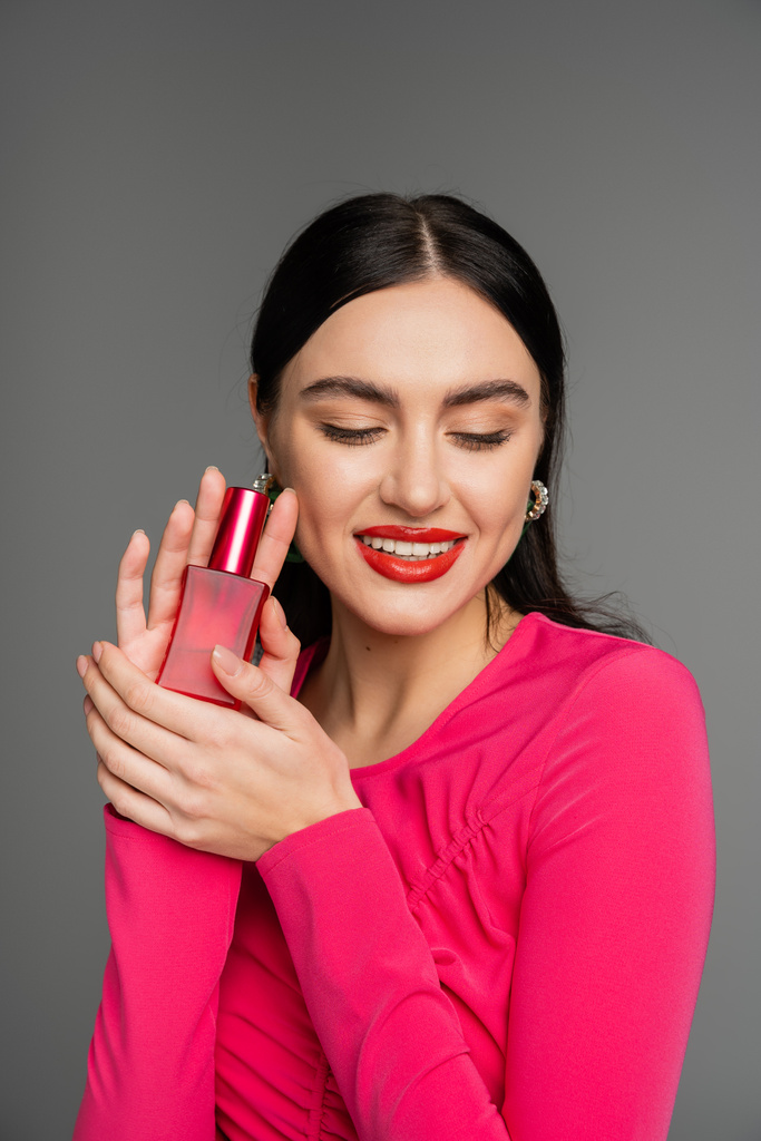retrato de una hermosa joven con cabello moreno y labios rojos posando en vestido magenta mientras sostiene la botella de perfume de lujo y sonriendo aislado sobre fondo gris  - Foto, imagen