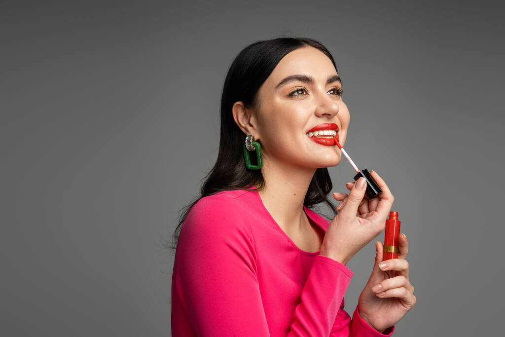 ευτυχισμένη γυναίκα με καστανά μαλλιά και μοντέρνα σκουλαρίκια εφαρμόζοντας κόκκινο lip gloss και χαμογελώντας ενώ ποζάρουν σε φούξια φόρεμα και την προετοιμασία για το κόμμα απομονώνονται σε γκρι φόντο  - Φωτογραφία, εικόνα