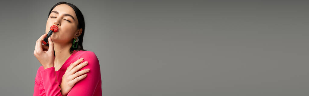 divertente donna con orecchini alla moda e capelli lucidi bruna applicare rossetto rosso e labbra pouting mentre posa isolata su sfondo grigio, banner  - Foto, immagini