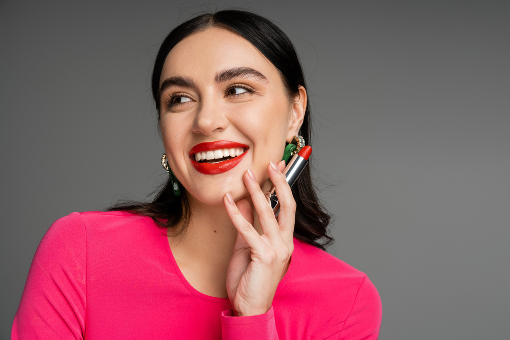 portrait of elegant woman with trendy earrings and shiny brunette hair holding red lipstick between fingers and smiling while posing on grey background  - Photo, Image