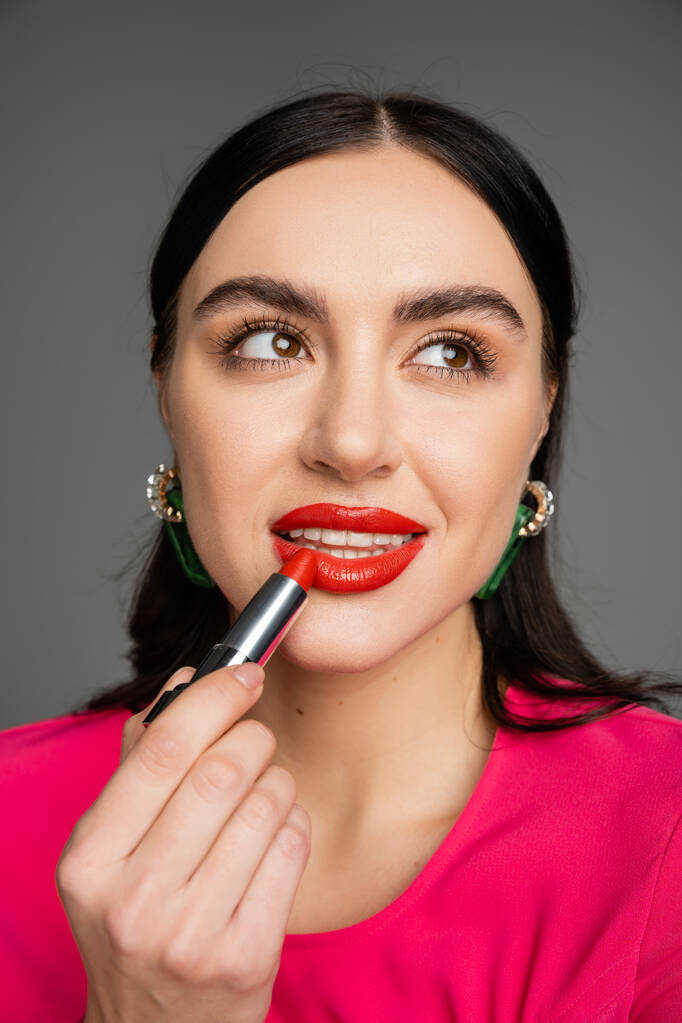 retrato de mujer joven impresionante con pendientes de moda y maquillaje impecable aplicando lápiz labial rojo mientras posando y mirando hacia otro lado sobre fondo gris  - Foto, imagen